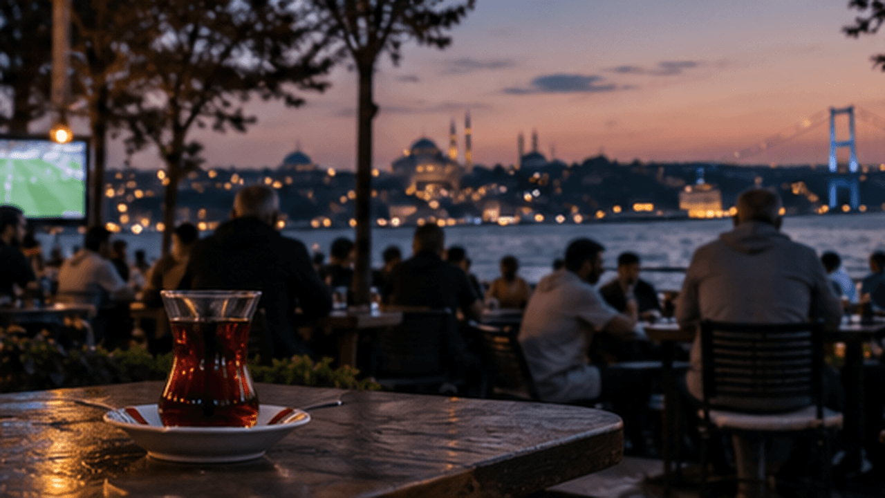 A glass of tea on a waterfront table at dusk, a football match glowing on a screen in the corner and the Bosphorus and a mosque skyline behind it