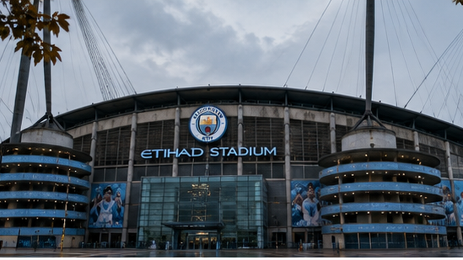 The Etihad Stadium exterior at dusk, floodlights on, the crest and stadium name lit above the entrance, the ground quiet during the international break
