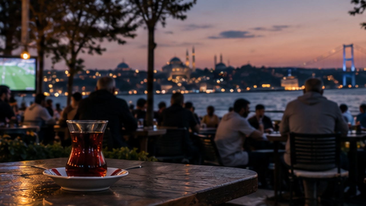A glass of tea on a waterfront table at dusk, a football match glowing on a screen in the corner and the Bosphorus and a mosque skyline behind it